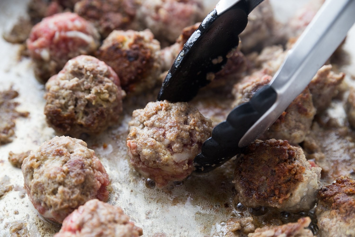 meatballs in a skillet with tongs flipping them 