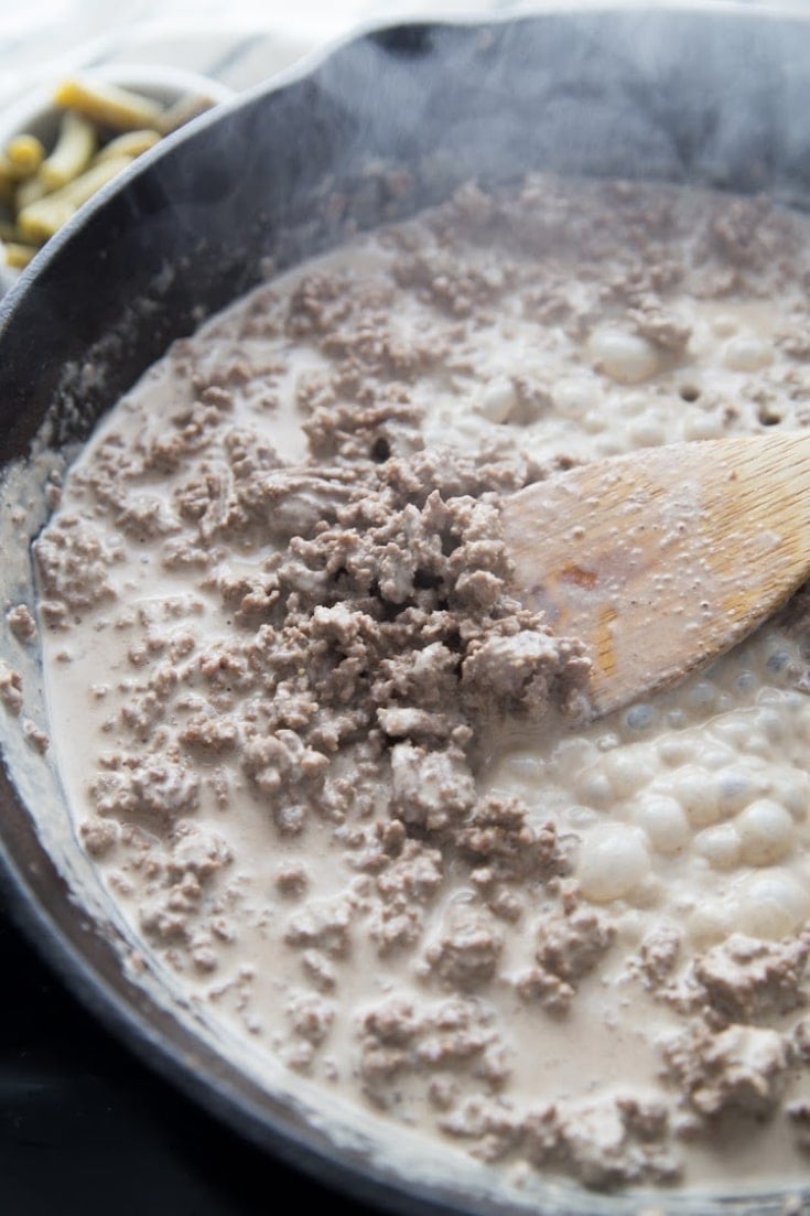 Overhead shot looking down at the thick and creamy keto ground beef casserole base featuring cooked crumbled beef and a creamy sauce with creaam cheese, heavy whipping cream, and beef broth simmering away in a black cast iron skillet, blended smoothly with a wooden spoon