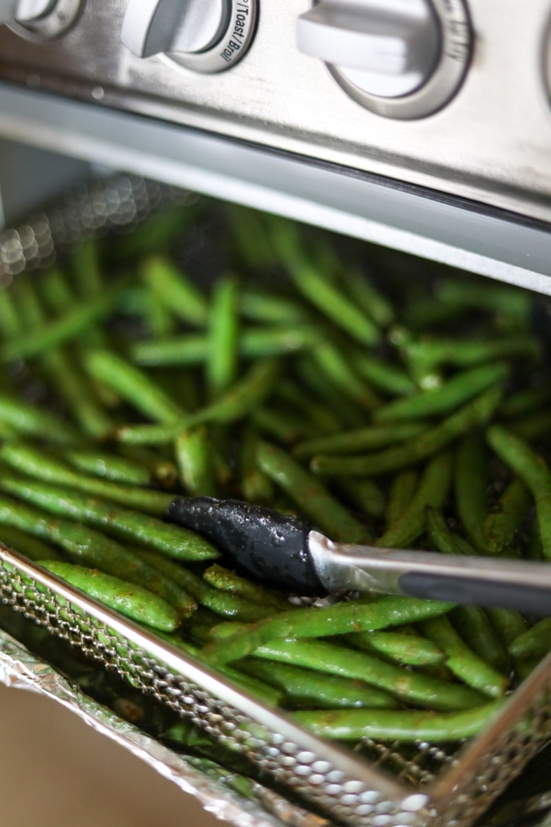 Green beans in air fryer basket with tongs.