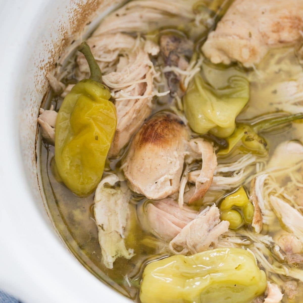 close shot of white crockpot with Mississippi chicken recipe with shredded chicken, broth and pepperocini peppers 
