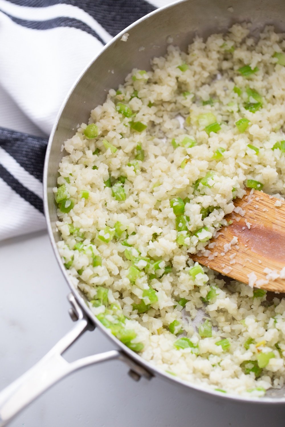 Riced cauliflower and chopped celery in a skillet with wooden spoon.