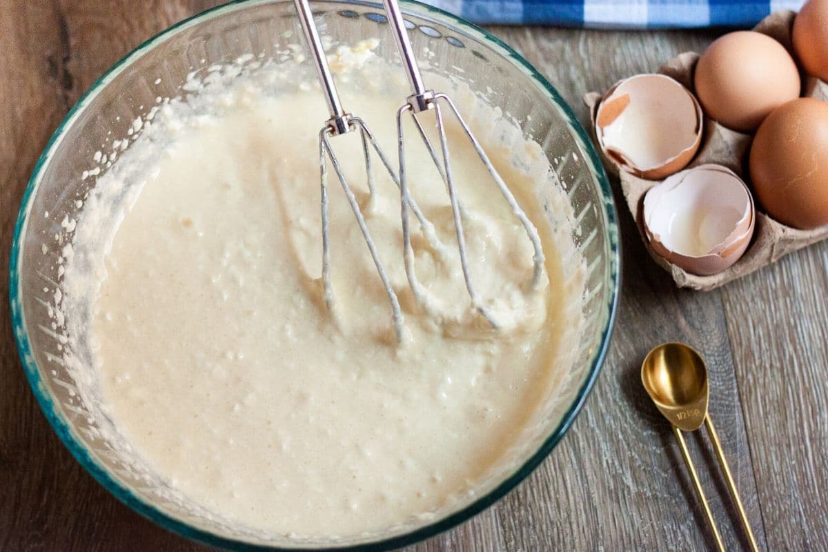 keto cheesecake batter in a clear bowl with beaters and cracked egg shells on table