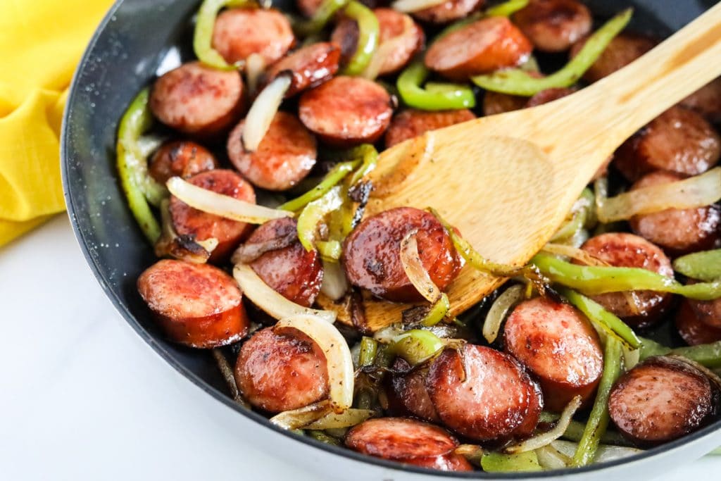 Overhead view of skillet filled with keto friendly smoked sausage, sliced bell peppers and onions cooked in olive oil. A wooden spoon dips into the skillet to serve out portions of the low carb sausage vegetable skillet dinner.