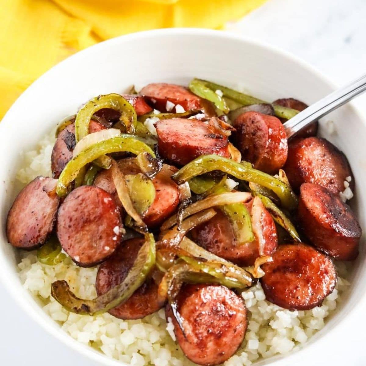 sausage with peppers and onions served over cauliflower rise in a white glass bowl.