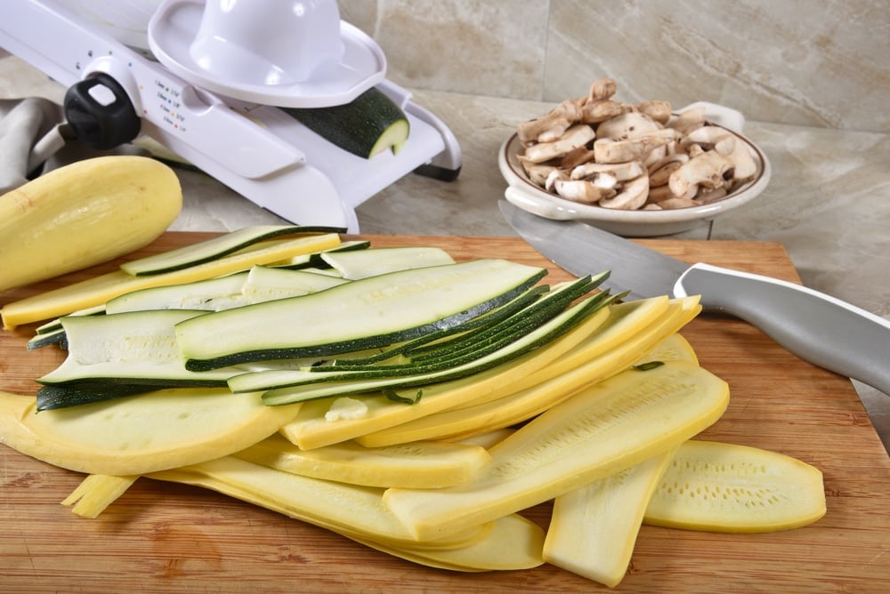 Ribbons of thin sliced zucchini and yellow squash on a cutting board with a mandolline slicer