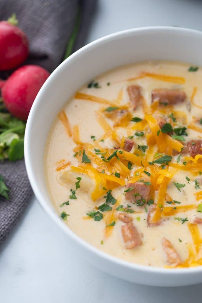 Close up photo looking down at bowl filled with creamy low carb cheesy smoked sausage soup made with sliced smoked sausage, cubed radishes, heavy cream, cheddar cheese, parsley and bacon bits on top for garnish