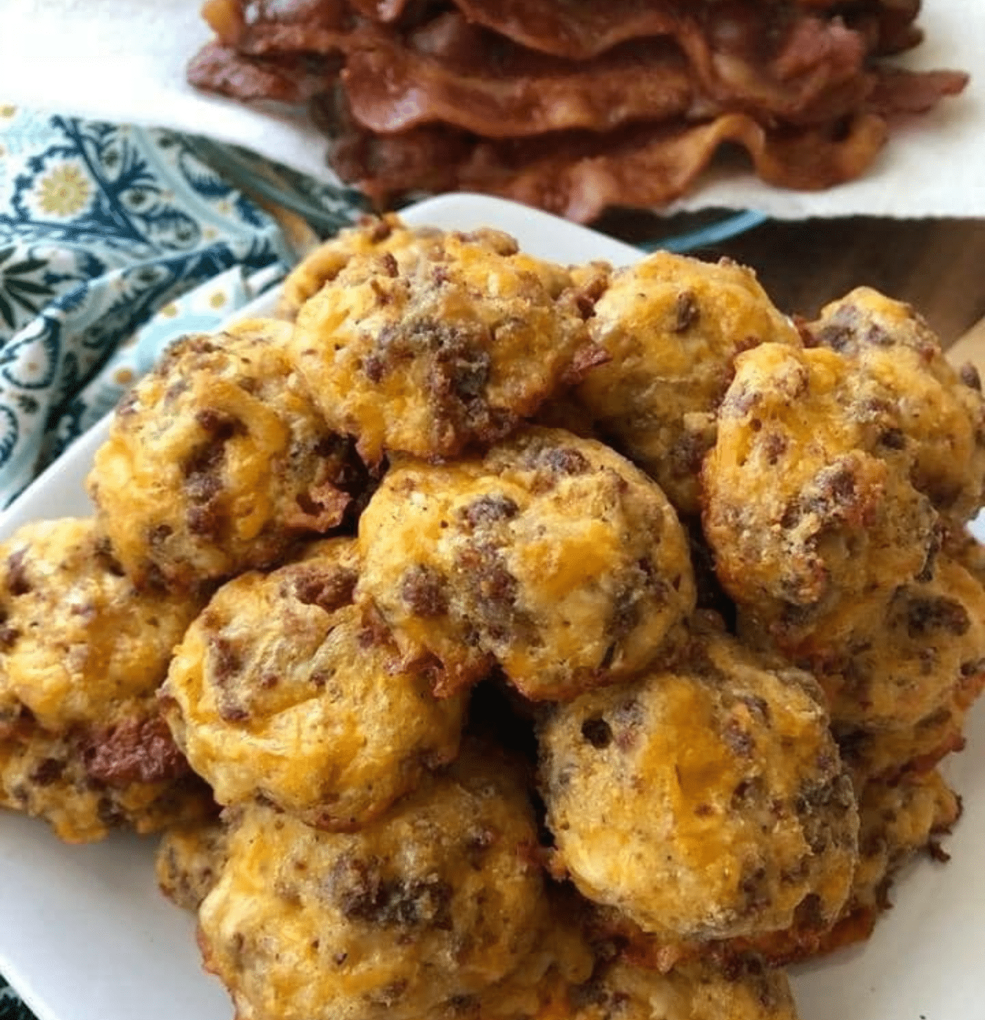 low-carb sausage balls piled on a white plate with bacon in thee background.
