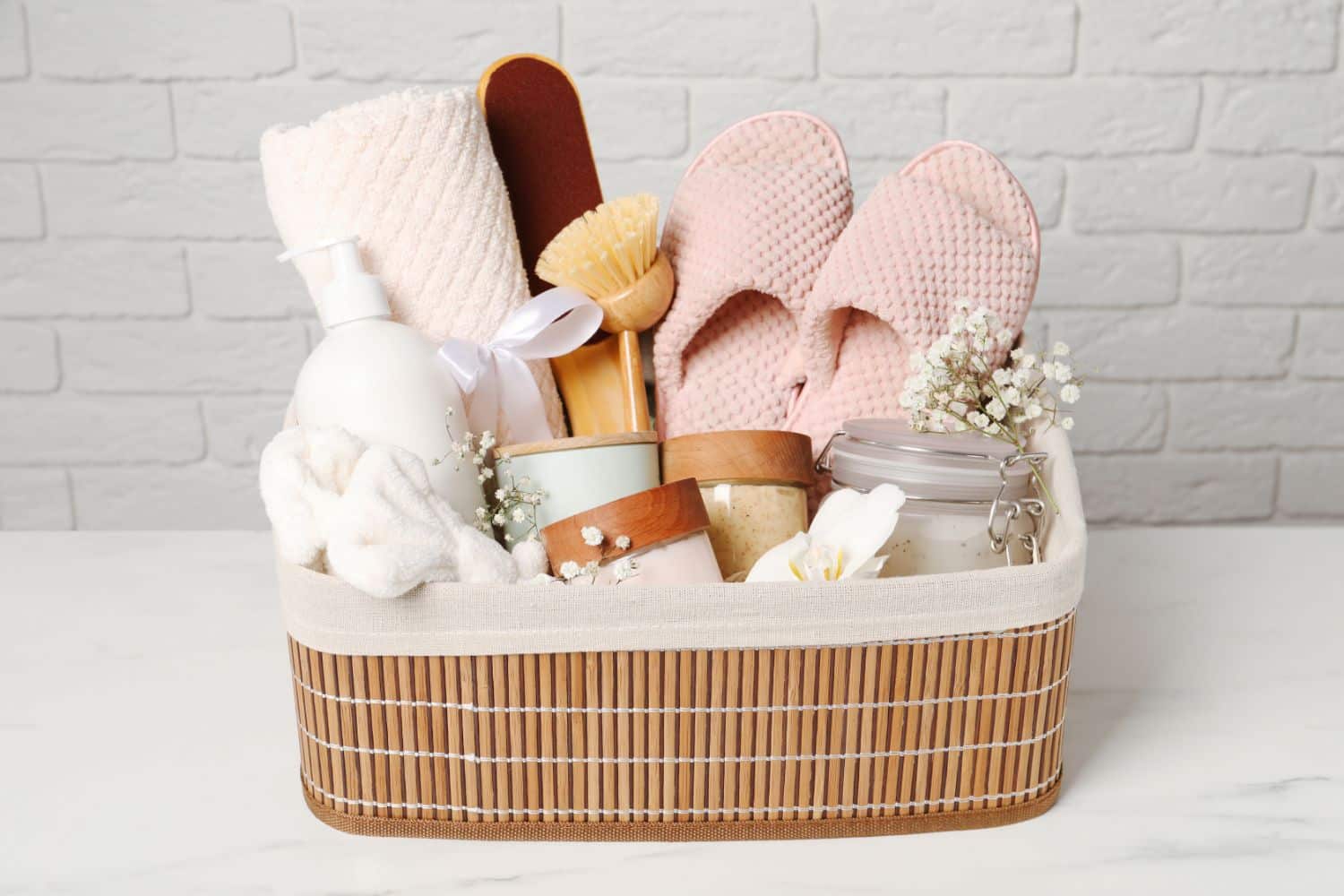 A wicker basket with a beige liner spa gift basket with spa products including slippers, candle, scrubbing brush, drying towel, candle, headband, salt scrub, and body wash