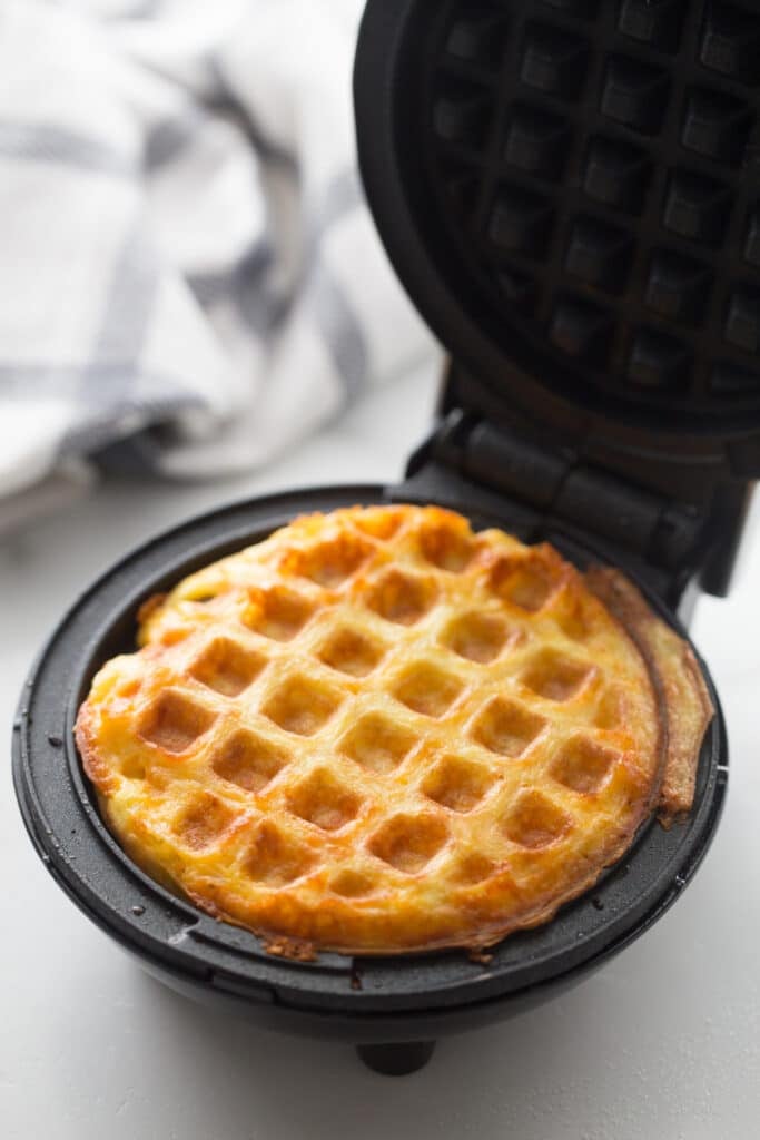 Close-up photo showcasing two circular golden brown pepperoni pizza chaffles actively baking inside an open mini dash waffle maker. The chaffles display a crispy hashbrown texture on the outside