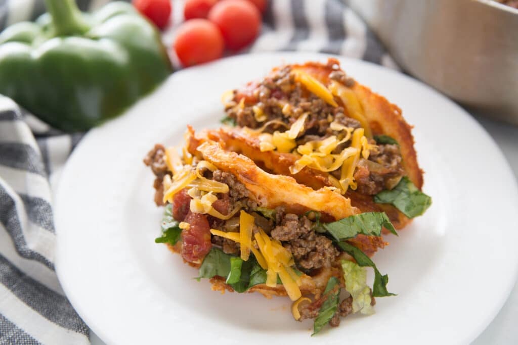 two taco meat stuffed chaffle taco shells on a white plate with cheese and tomato toppings