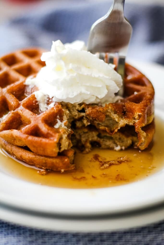 Two round golden brown pumpkin chaffles stacked high on white plates drizzled with syrup and topped with whipped cream, fork inserted into front chaffle with piece sliced to show fluffy interior texture