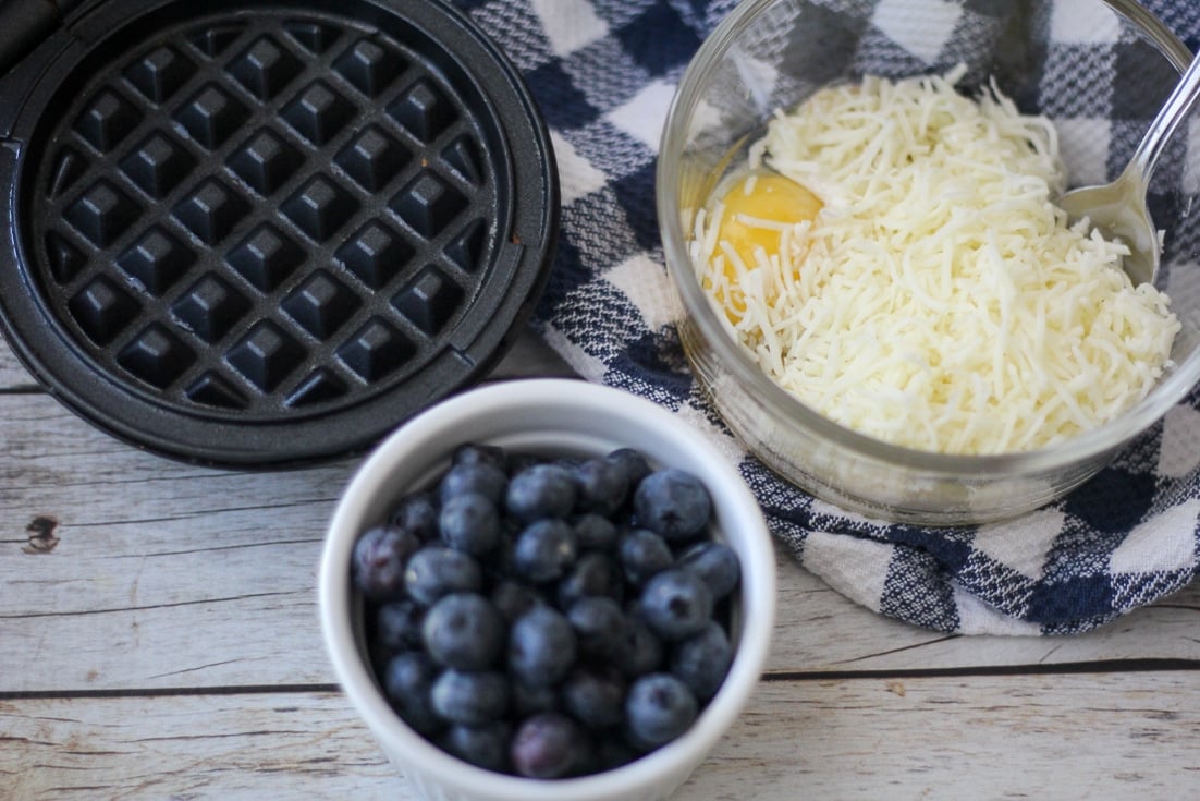 Dash mini waffle maker open on left; small ramekin filled with blueberries in center; glass bowl on right filled with separated ingredients for blueberry chaffle batter including egg, shredded cheese, cream cheese, coconut flour, baking powder, vanilla, and cinnamon