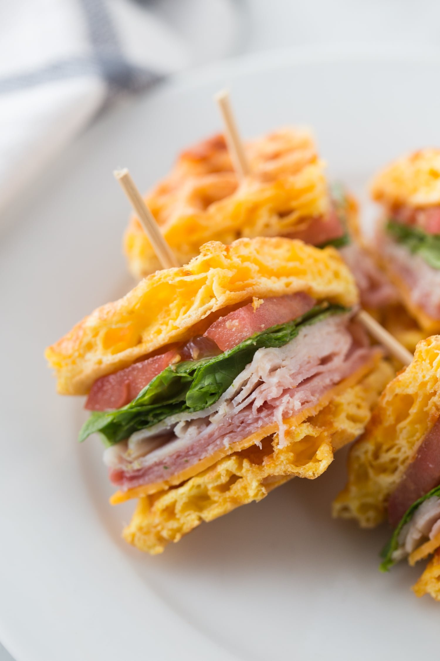 Close-up photo of a quarter of a chaffle club sandwich on a white plate, featuring a chaffle bun filled with turkey, ham, tomato, lettuce, and toothpicks holding it together and the other quarters in the background, 