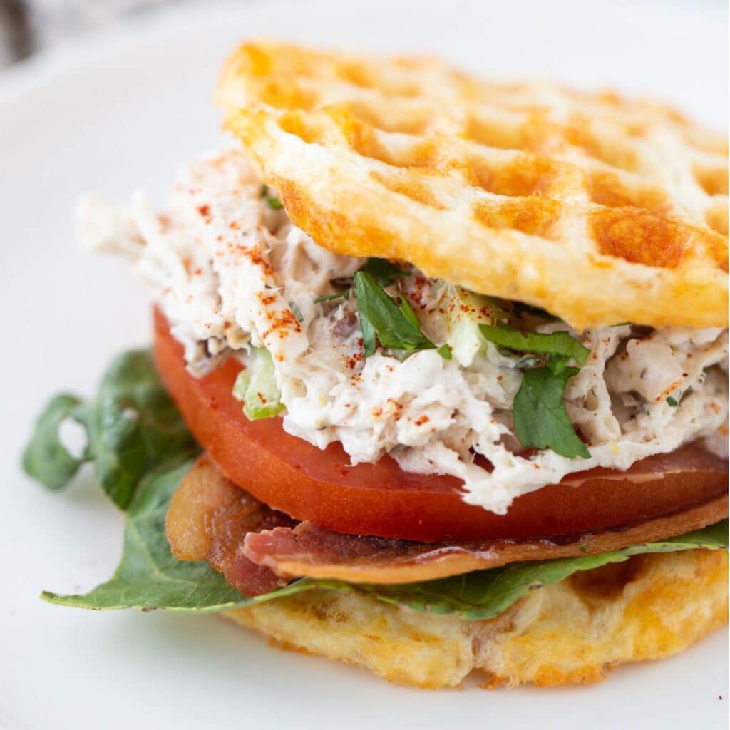 Square close-up photo of keto chicken salad sandwich served in chaffle buns topped with lettuce leaves and tomato slice
