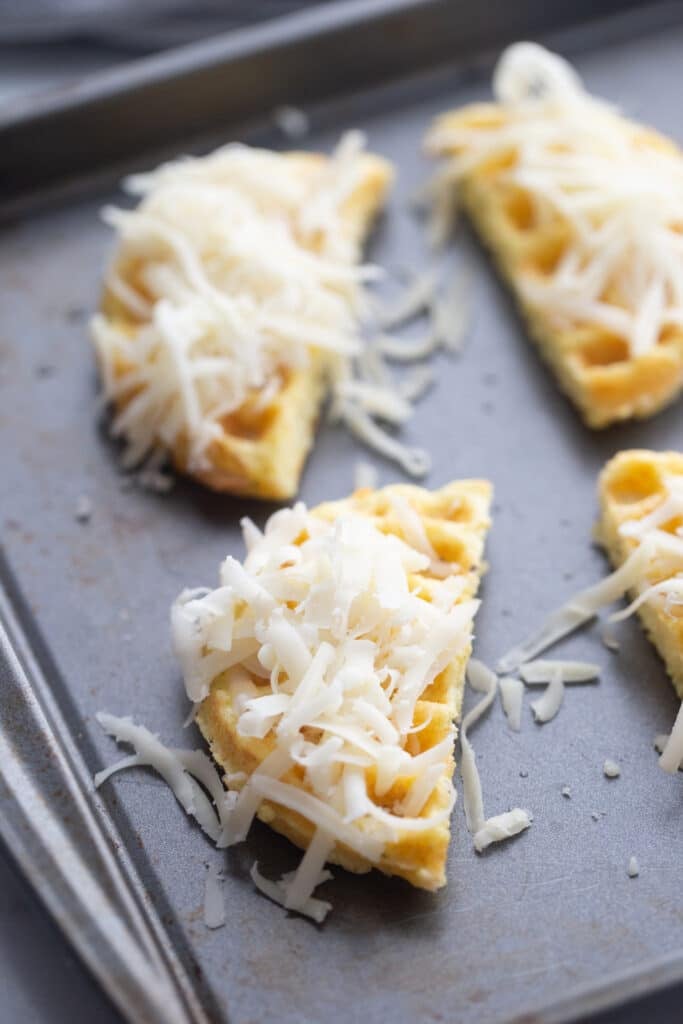 Several sliced chaffle rounds arranged in a single layer on a large silver baking sheet covered evenly with freshly grated mozzarella cheese atop the crisp garlic bread before putting in the hot oven where the cheese will gently melt into delicious gooey bites.