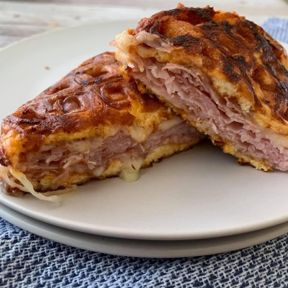 Hot ham and cheese chaffle sandwich cut in half diagnolly and stacked on top of each other so you can see the melty cheese. It is a round plate that is stacked on another round plate with a blue kitchen towel towards the front of the picture.