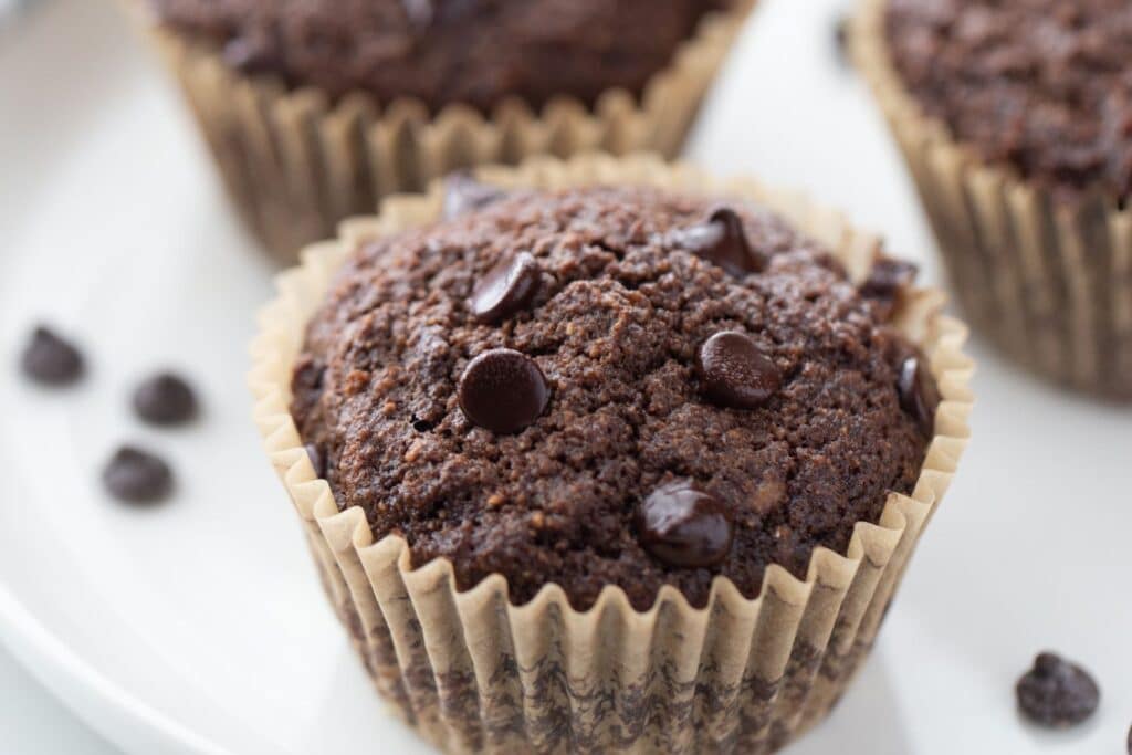 3 double chocolate muffins in a triangle angled shot on a white plate with chocolate chips sprinkled around it 
