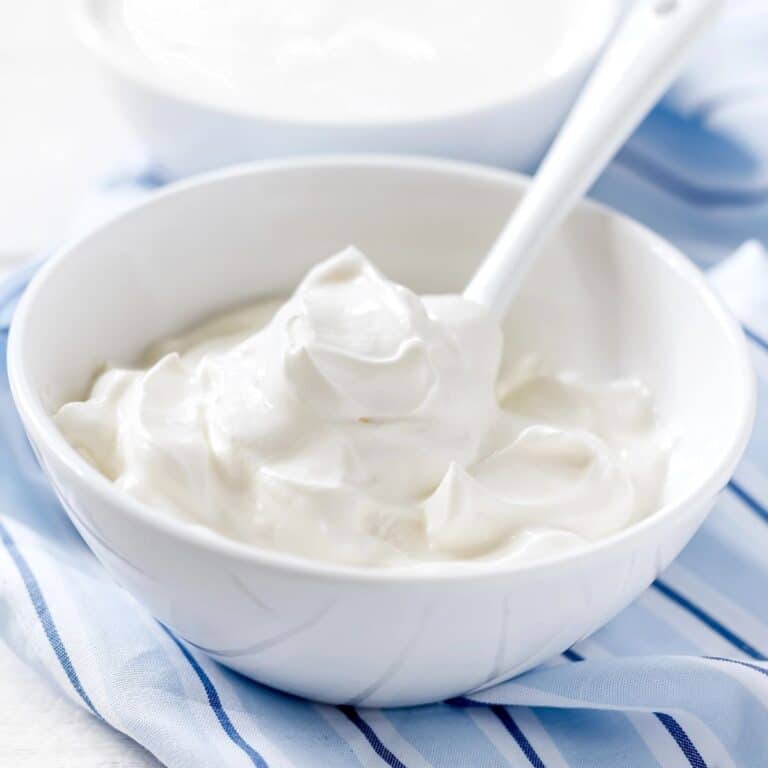 Close-up of a white bowl filled with creamy sour cream, a white spoon, and a blue and white napkin behind it, angled shot