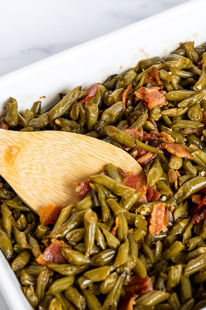 Stirring the crack green beans casserole with a wooden spoon to evenly distribute the flavorful bacon and seasoning mixture throughout the dish.