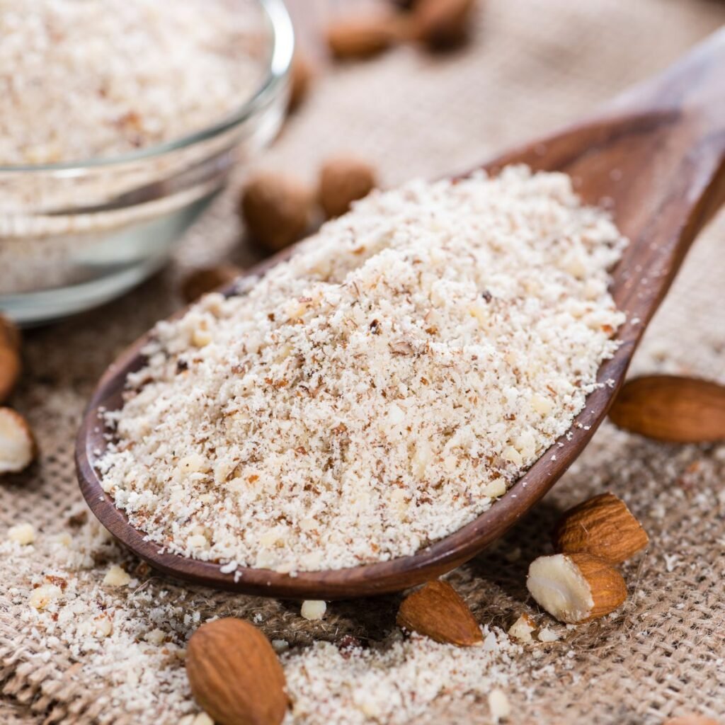 A close-up of a wooden spoon filled with almond flour, surrounded by whole almonds, a glass bowl of almond flour, and loose flour on a countertop, illustrating the concept of almond flour and its substitutes in baking and cooking.