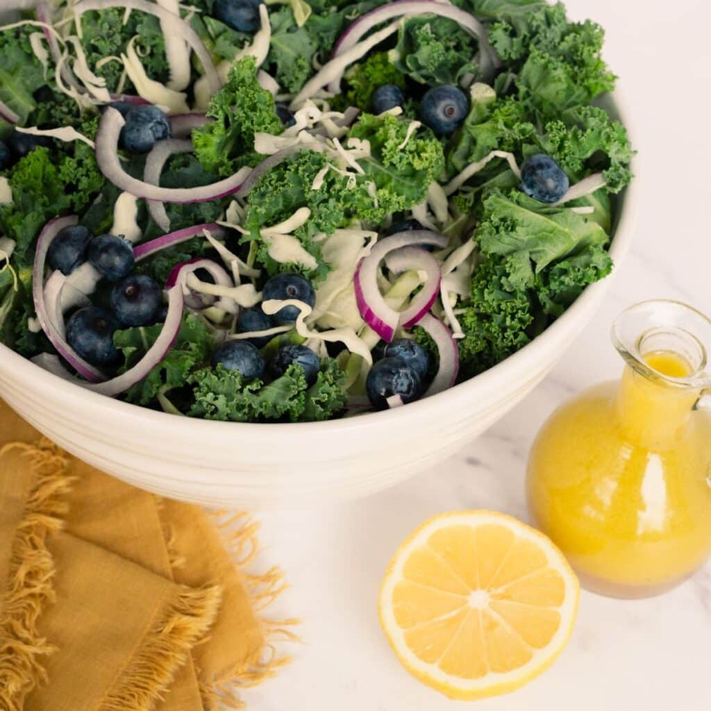 A square image featuring the visually stunning Kale Crunch Salad in a white bowl, set against a crisp white background, with a pop of color from the vibrant yellow cloth and fresh lemon slices, emphasizing the salad's beautiful presentation and tantalizing appeal.