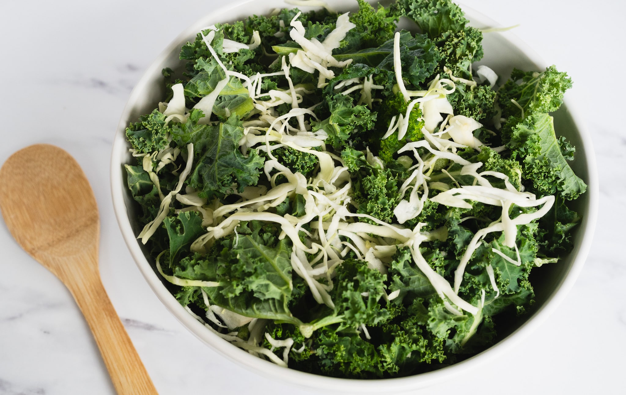 A process image showing a mixture of chopped kale and shredded cabbage in a white bowl, with a wooden spoon placed next to it, set against a clean white background, illustrating a step in the preparation of the Kale Crunch Salad recipe.
