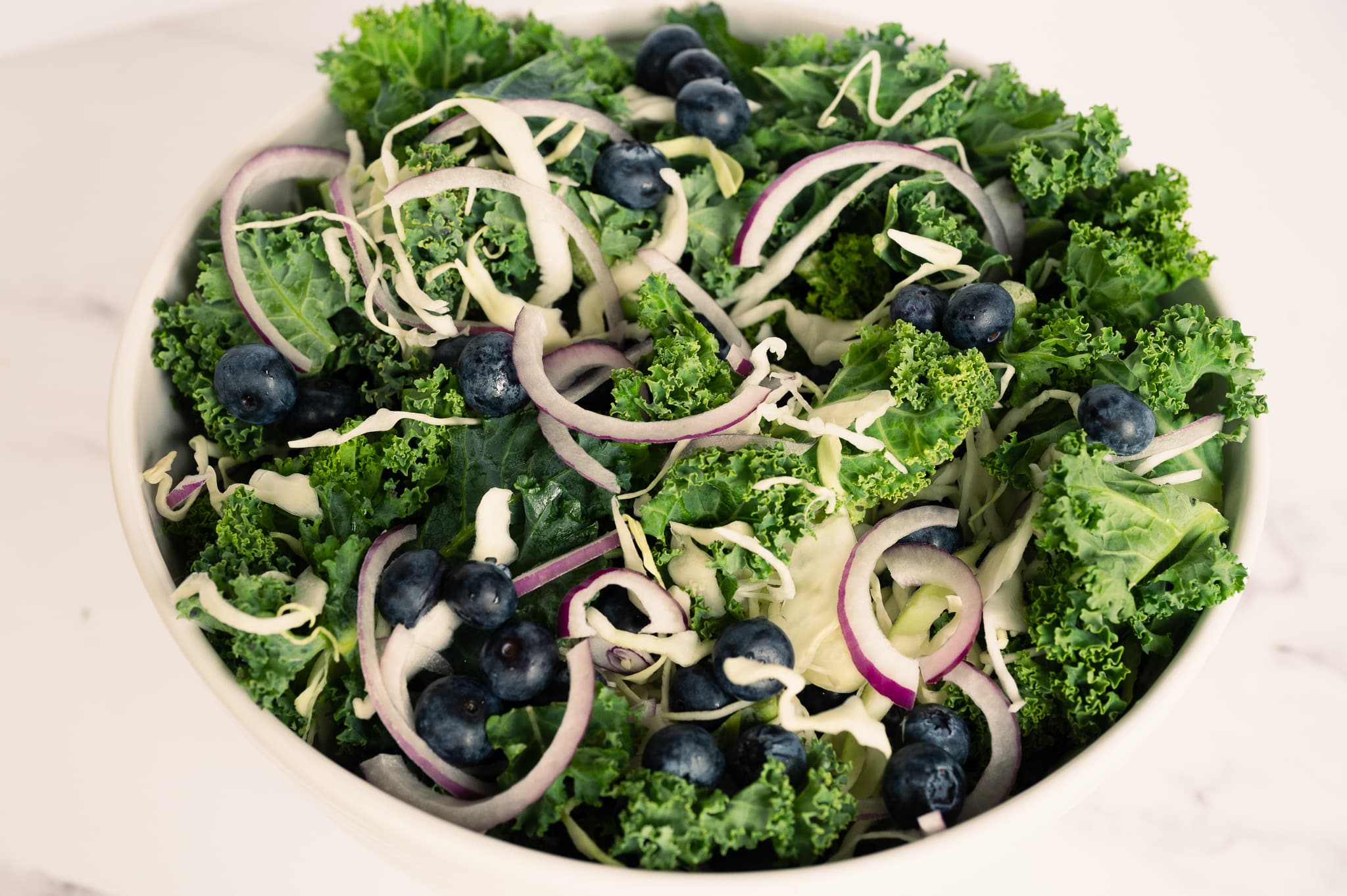 A process image depicting the Kale Crunch Salad mixture of chopped kale, shredded cabbage, fresh blueberries, and sliced red onions in a white bowl, with a wooden spoon nearby, set against a white background, showcasing the colorful ingredients being combined in the salad preparation.