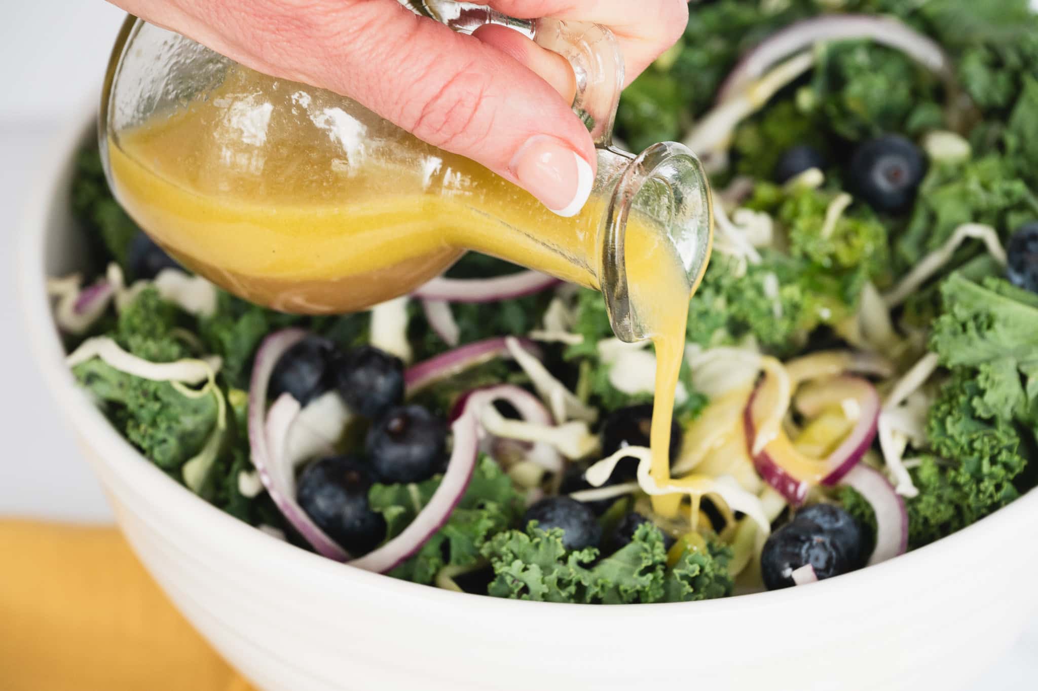 A close-up image of the completed Kale Crunch Salad in a white bowl, featuring a mixture of kale, cabbage, blueberries, red onions, and toasted nuts, with a stream of Apple Cider and Dijon Mustard vinaigrette being poured over the top, highlighting the final step in preparing this delicious and healthy salad.