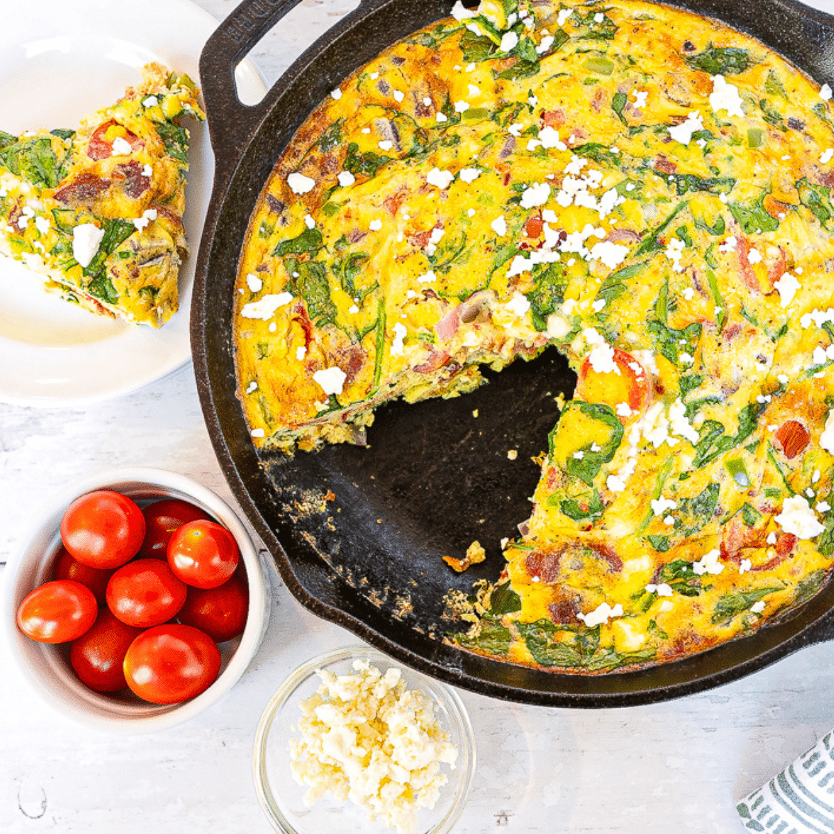 Keto bacon and vegetable frittata in a black skillet with a slice removed, served alongside a small baking dish with cherry tomatoes and cheese, on a white background.