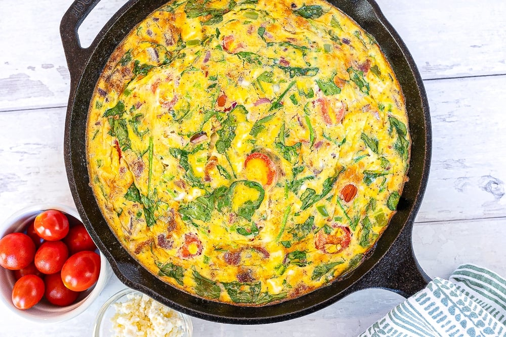 A finished keto bacon and vegetable frittata baked to golden perfection in a black skillet, set against a white background.