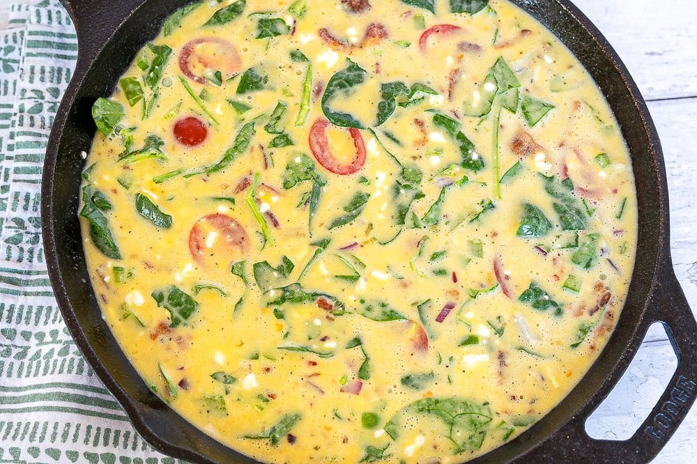 A process image showing the mixed keto frittata egg and vegetable mixture being poured into a black baking skillet, set against a white background.