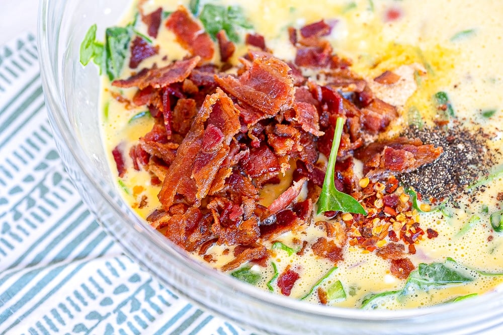 A process image displaying the addition of crispy bacon pieces to the vegetable and egg mixture for a keto frittata, using a whisk in a clear baking bowl, set against a white background.