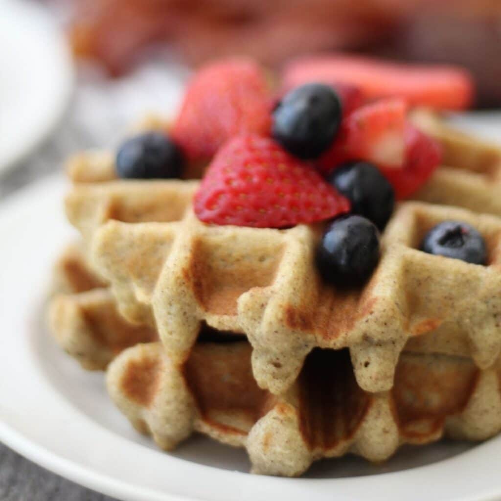 Alt text: Close-up of golden brown waffles topped with fresh strawberries and blueberries on a white plate. The waffles appear to have a slightly grainy texture, since they are keto-friendly.
