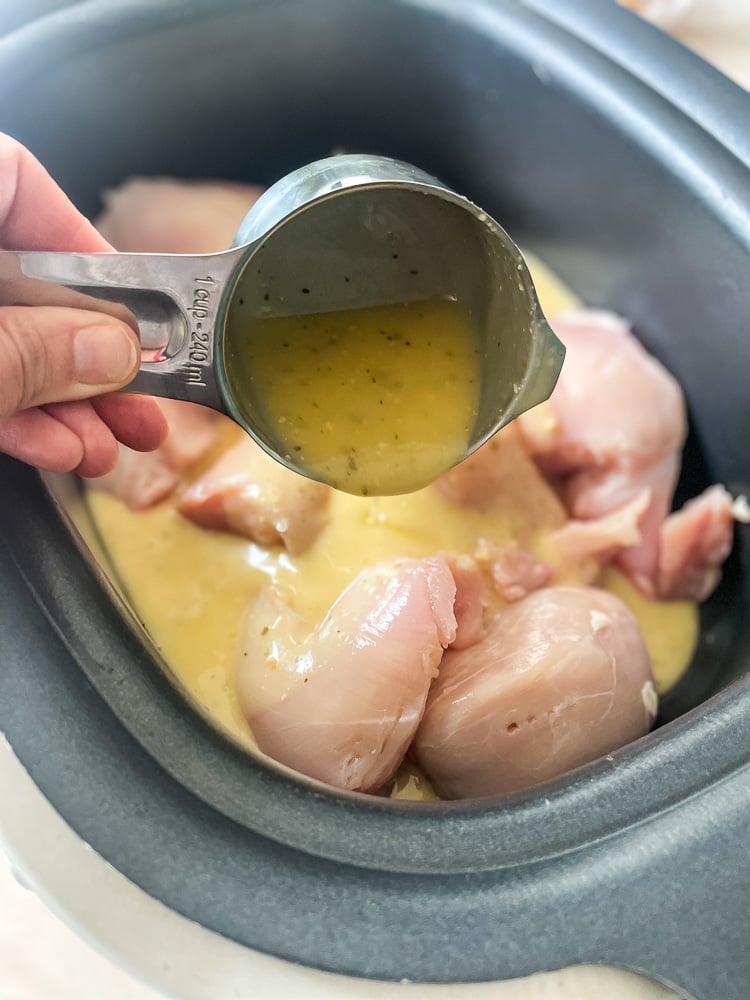 Raw chicken breasts in a slow cooker with Olive Garden dressing being poured from a measuring cup, showing the first step of preparing the viral crock pot chicken recipe