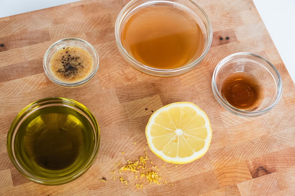 Ingredients for cider vinaigrette dressing on a wooden cutting board: bowls of olive oil, apple cider vinegar, honey or maple syrup, Dijon mustard, and a halved lemon with lemon zest. Black pepper visible in one bowl