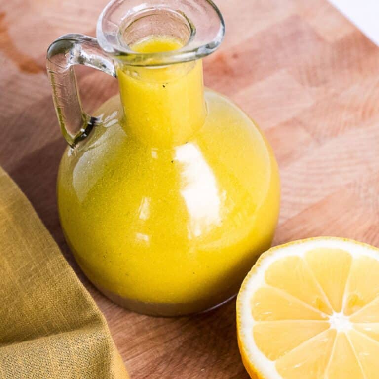 Glass bottle of homemade cider vinaigrette dressing with a bright yellow color, placed on a wooden surface next to a halved lemon and a folded mustard-colored napkin
