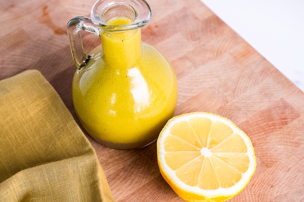 Glass bottle of homemade cider vinaigrette dressing with a bright yellow color, placed on a wooden surface next to a halved lemon and a folded mustard-colored napkin