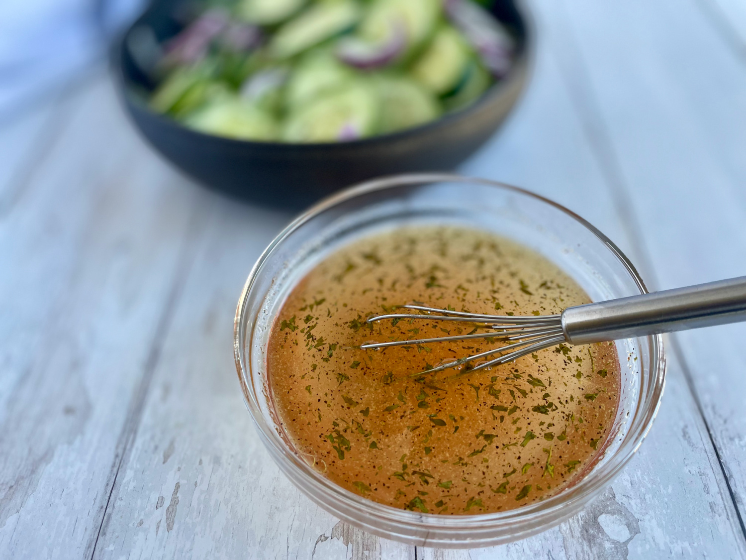 A black bowl filled with sliced cucumbers and red onions, with a small clear bowl in front containing a whisk and a tangy vinegar-based dressing, ready to mix for a keto cucumber onion salad.