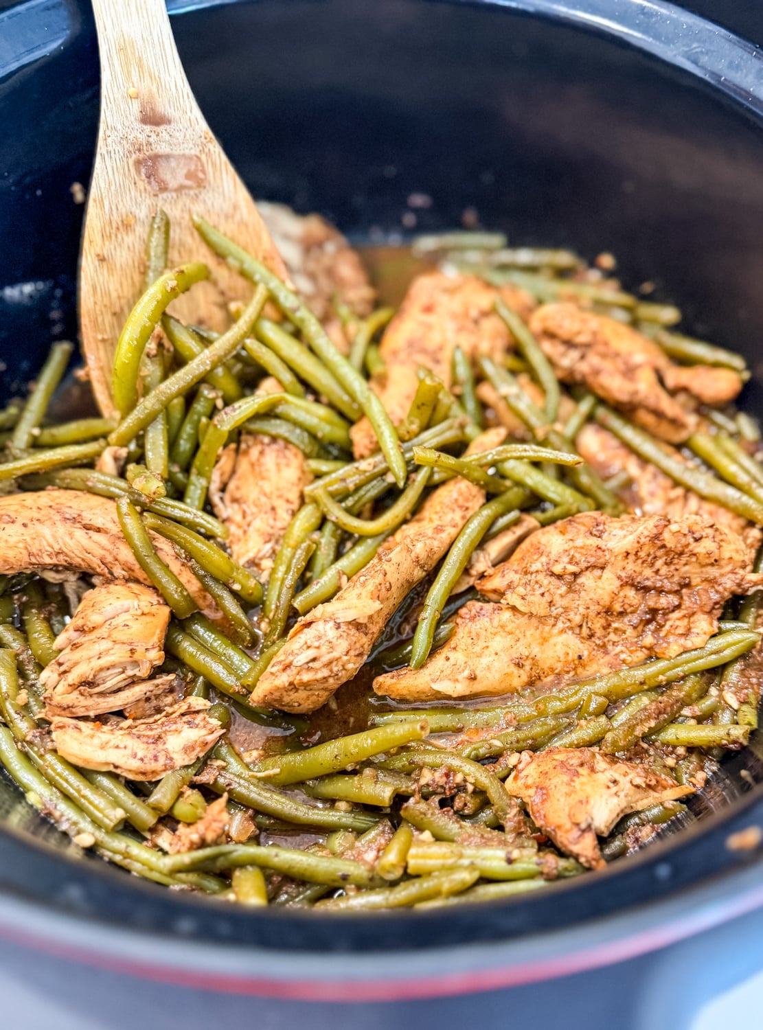 Juicy chicken tenders and green beans cooked in honey garlic sauce inside a crockpot, stirred with a wooden spoon.